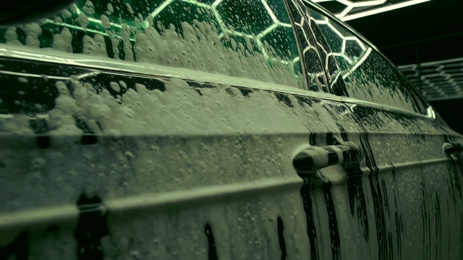 Thick white foam sliding down the black flank of a car, green honeycomb lights reflected in the wet paint.