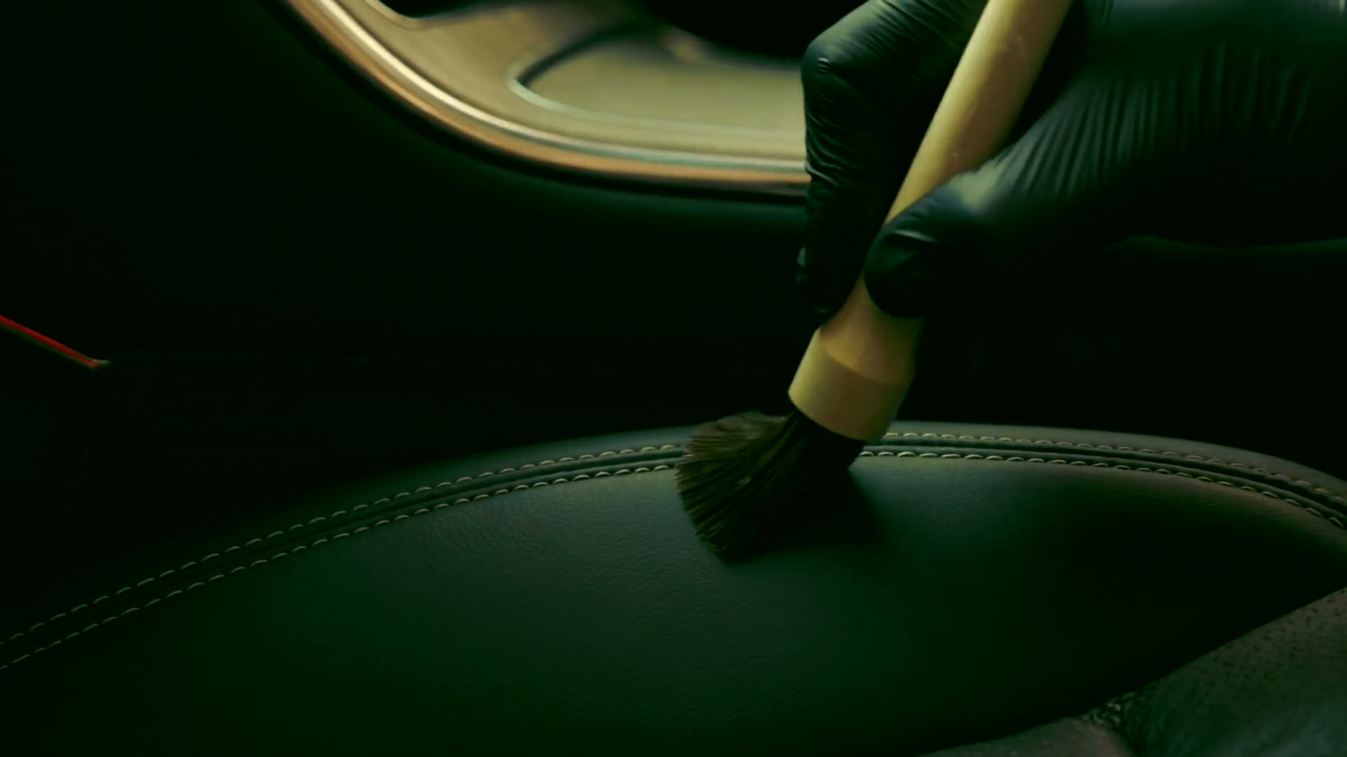 A gloved hand working a soft detailing brush along the stitching of a black leather car seat.
