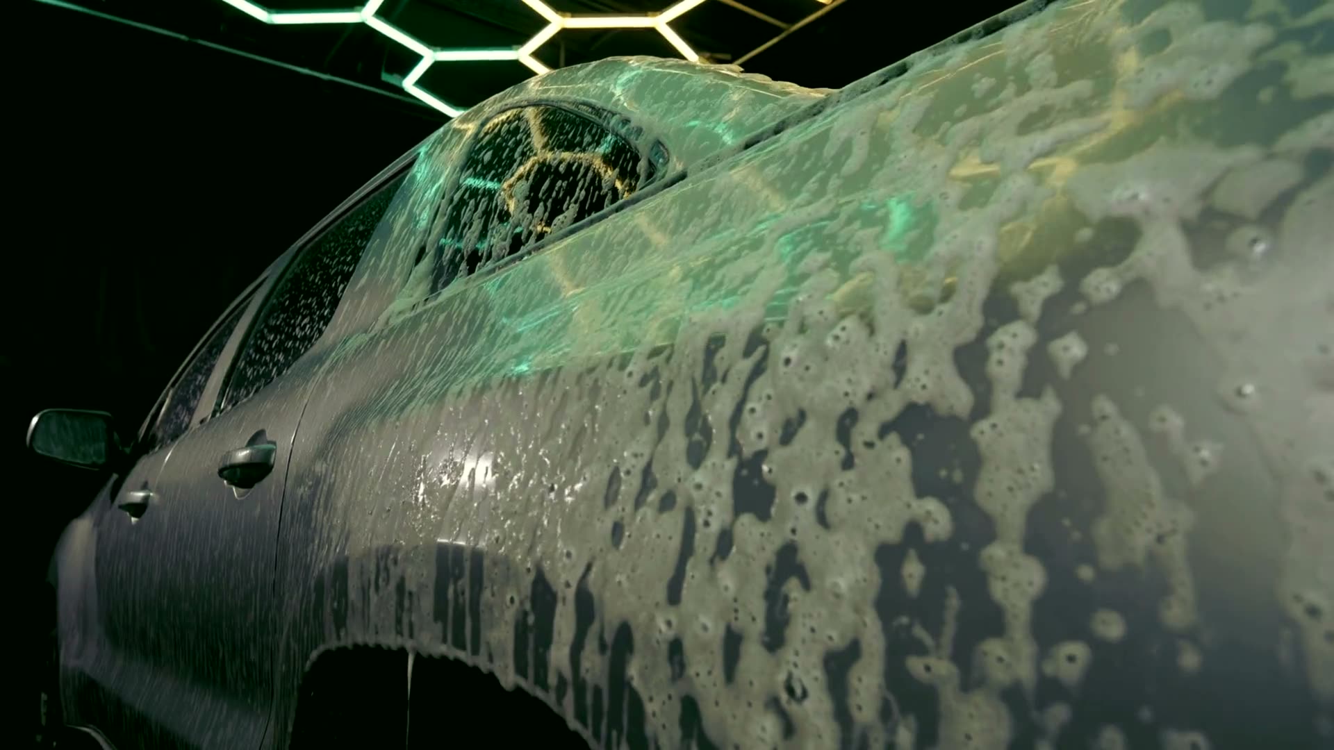 Thick foam sliding down the large flank of a dark vehicle, green honeycomb light reflected through the suds.