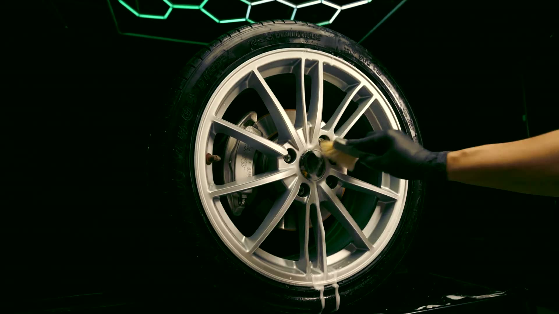 A gloved hand brushing between the spokes of an alloy wheel, suds running off the polished faces.